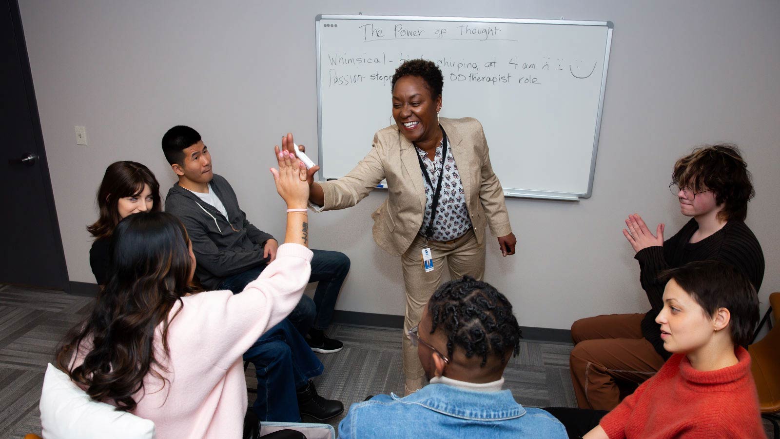 A therapist gives a high five to a participant in a group session, where other attendees are seated in a circle in a room with a whiteboard titled "The Power of Thought".