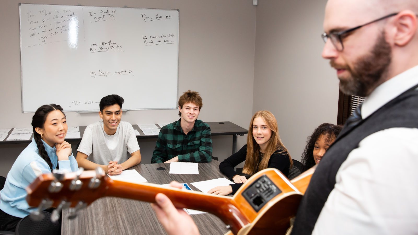 A music therapy session with a group of young people smiling and paying attention to a person holding a guitar.