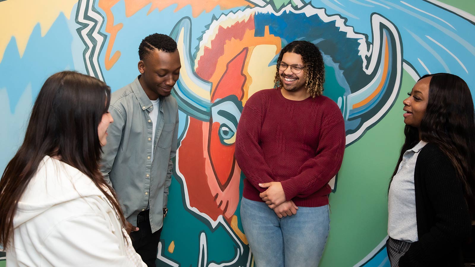 A small group of young adults laughing together in front of a colorful mural.