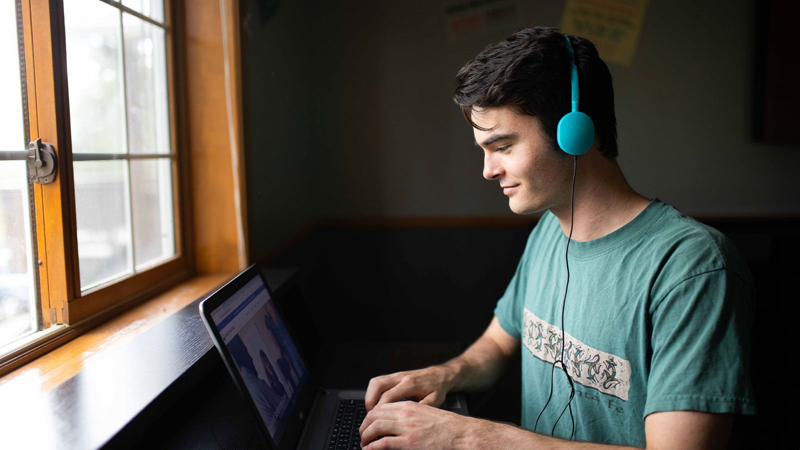 A young man with headphones working on a laptop by a window.