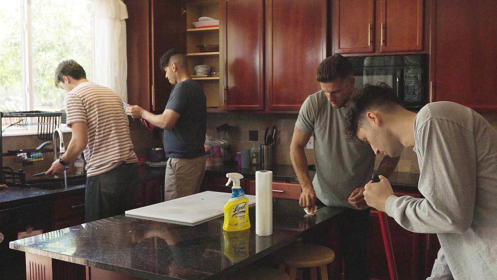 Several men participating in a kitchen cleanup, wiping down countertops and washing dishes.
