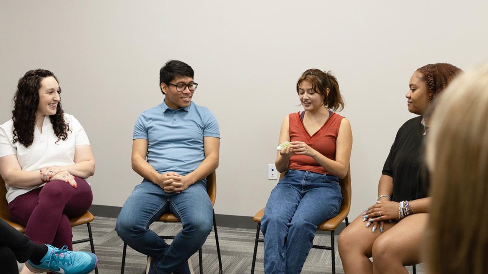 A circle of people seated in group therapy, smiling and interacting with each other.