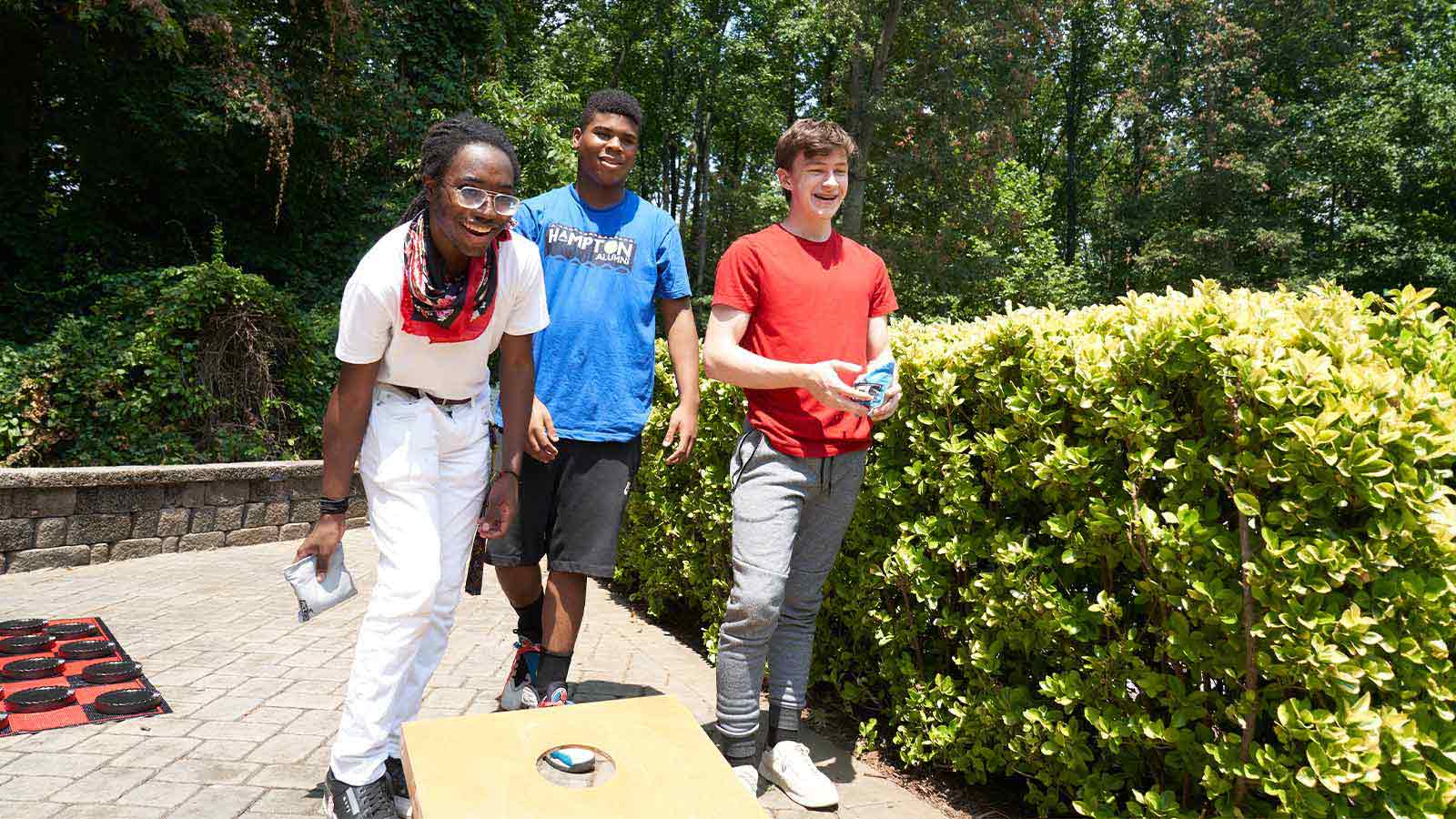 Young adults playing a bean bag toss game on a sunny day outdoors.