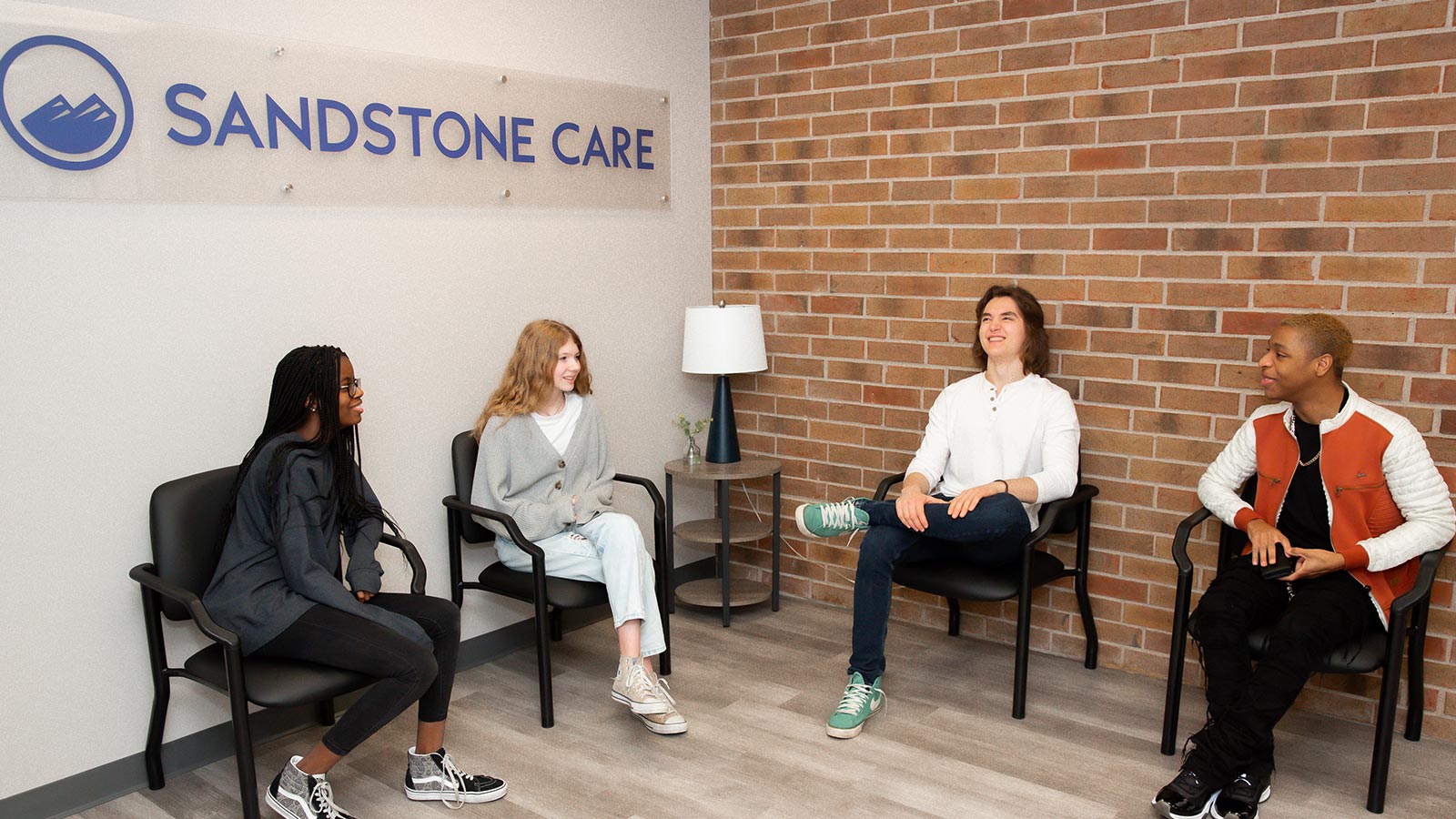 Four people sitting in a waiting area with a sign saying "Sandstone Care," having a relaxed conversation.