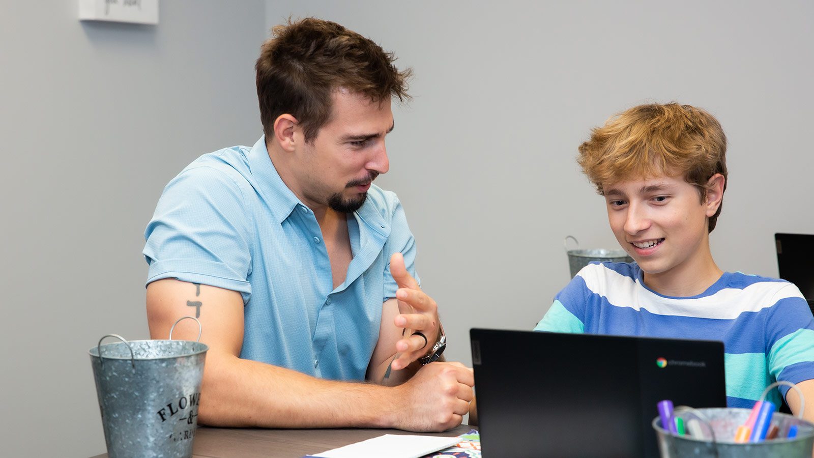 A man and teenage boy engaging in a one-on-one conversation across a desk with a laptop.