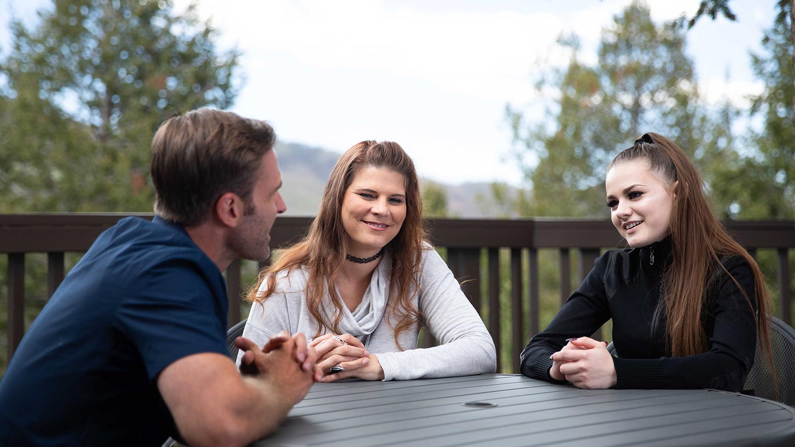 Three people engaged in a friendly discussion at an outdoor table, surrounded by a tranquil natural landscape.