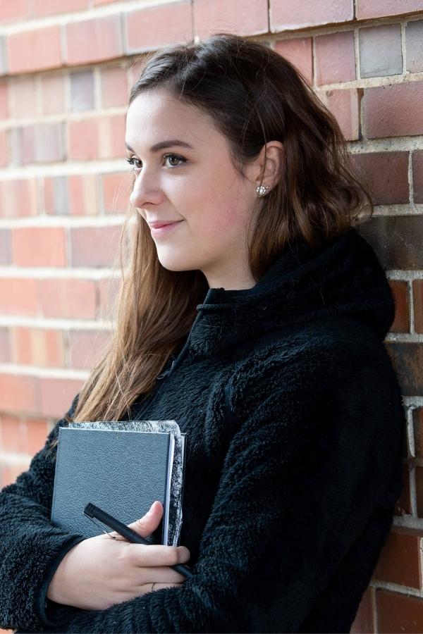 A teenage girl standing with a notebook and smiling.