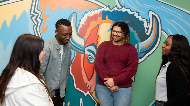 A group of four young adults are chatting and laughing together in front of a colorful, abstract mural.