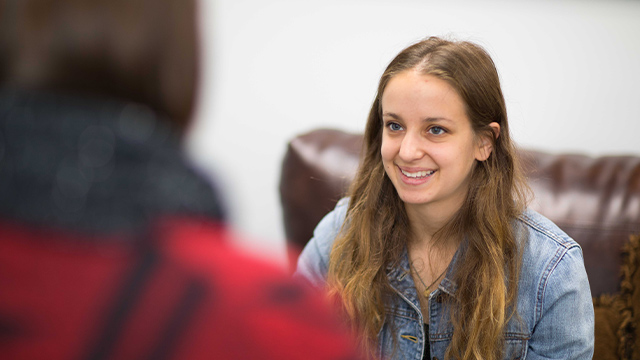 A young woman in a denim jacket smiles warmly during a conversation.