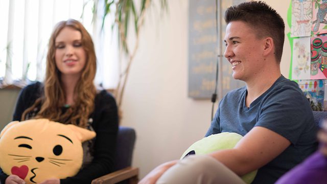 Two young adults sit smiling, holding playful cushions, in a room with colorful artwork on the wall.