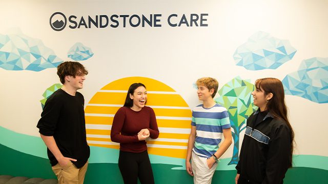 Four young adults are laughing and chatting in front of a colorful mural with the Sandstone Care logo.