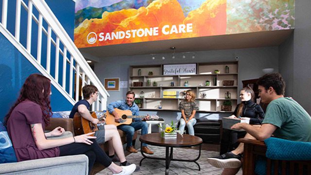Young adults enjoy a music session with a guitar in a cozy living room adorned with a vibrant mural.