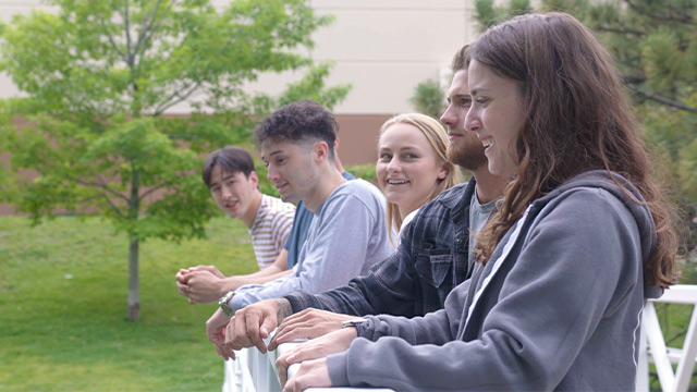 A group of young adults leans on a balcony railing, laughing and enjoying the outdoor setting together.