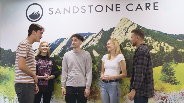 Five young adults are standing and engaging with each other in a friendly manner, with a mountain landscape mural and the Sandstone Care logo above them.