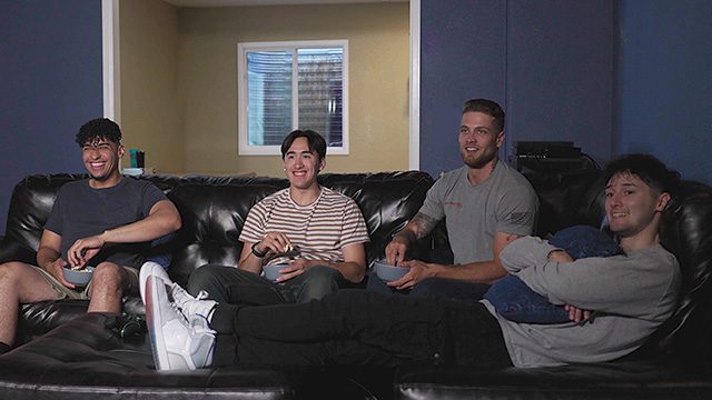 Four young men are laughing and relaxing on a large leather couch in a home theater setting.
