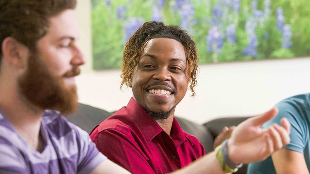 A man with a beaming smile talks with his peers in a relaxed living room environment.