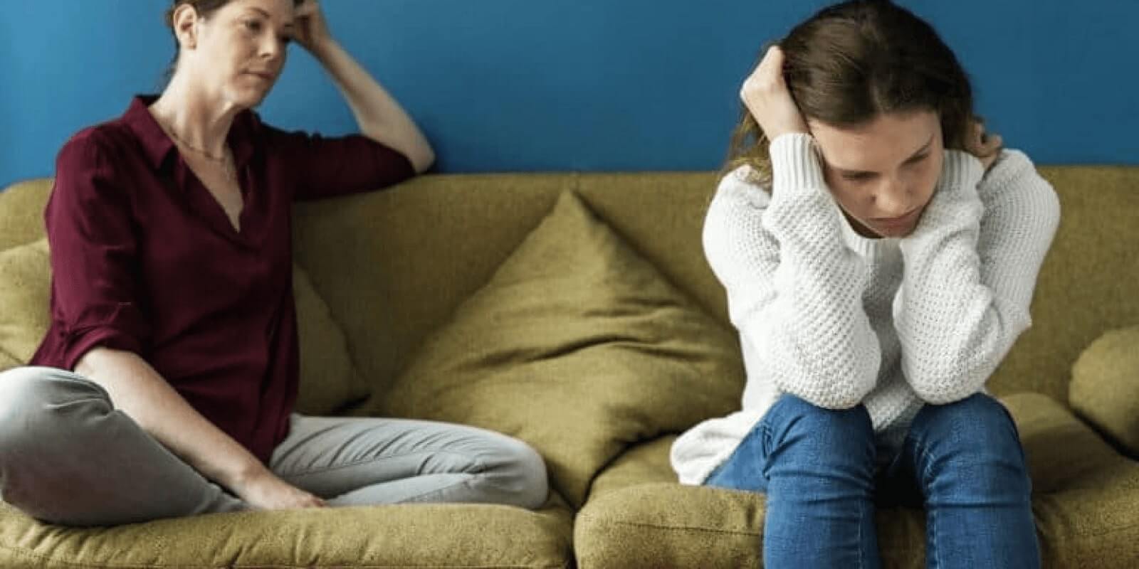 A teen holding her head looking depressed while her mom looks at her on the same couch