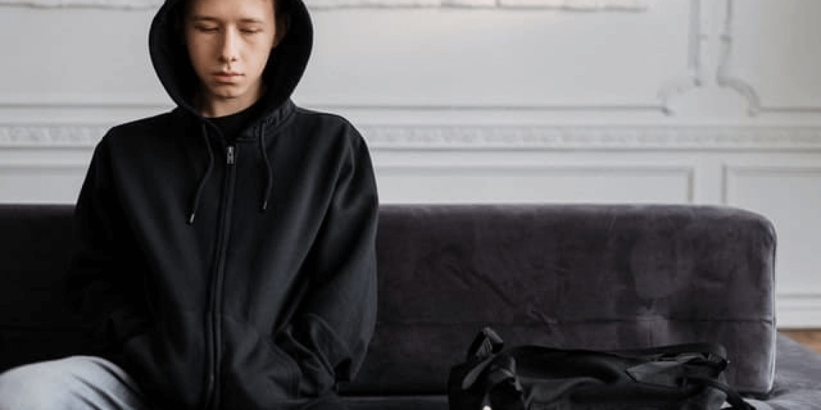 A teen boy wearing a black hoodie looking down while sitting on a sofa with his backpack