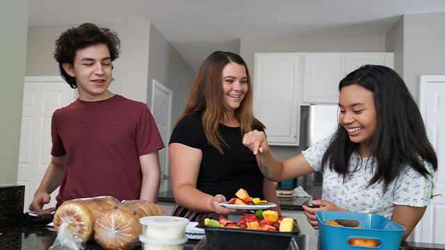 Three teenagers happily engage in preparing food together in a bright kitchen.