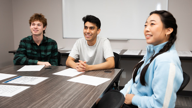 Three young adults smile and sit attentively at a table with papers in a classroom setting.