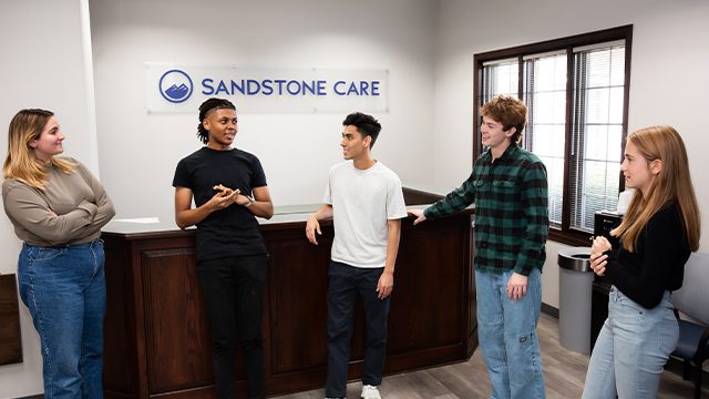 Five young adults stand and chat near a reception desk with the logo "Sandstone Care" on the wall in a bright office setting.