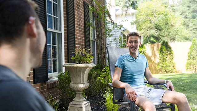 A cheerful young man converses in an outdoor seating area by a brick house.