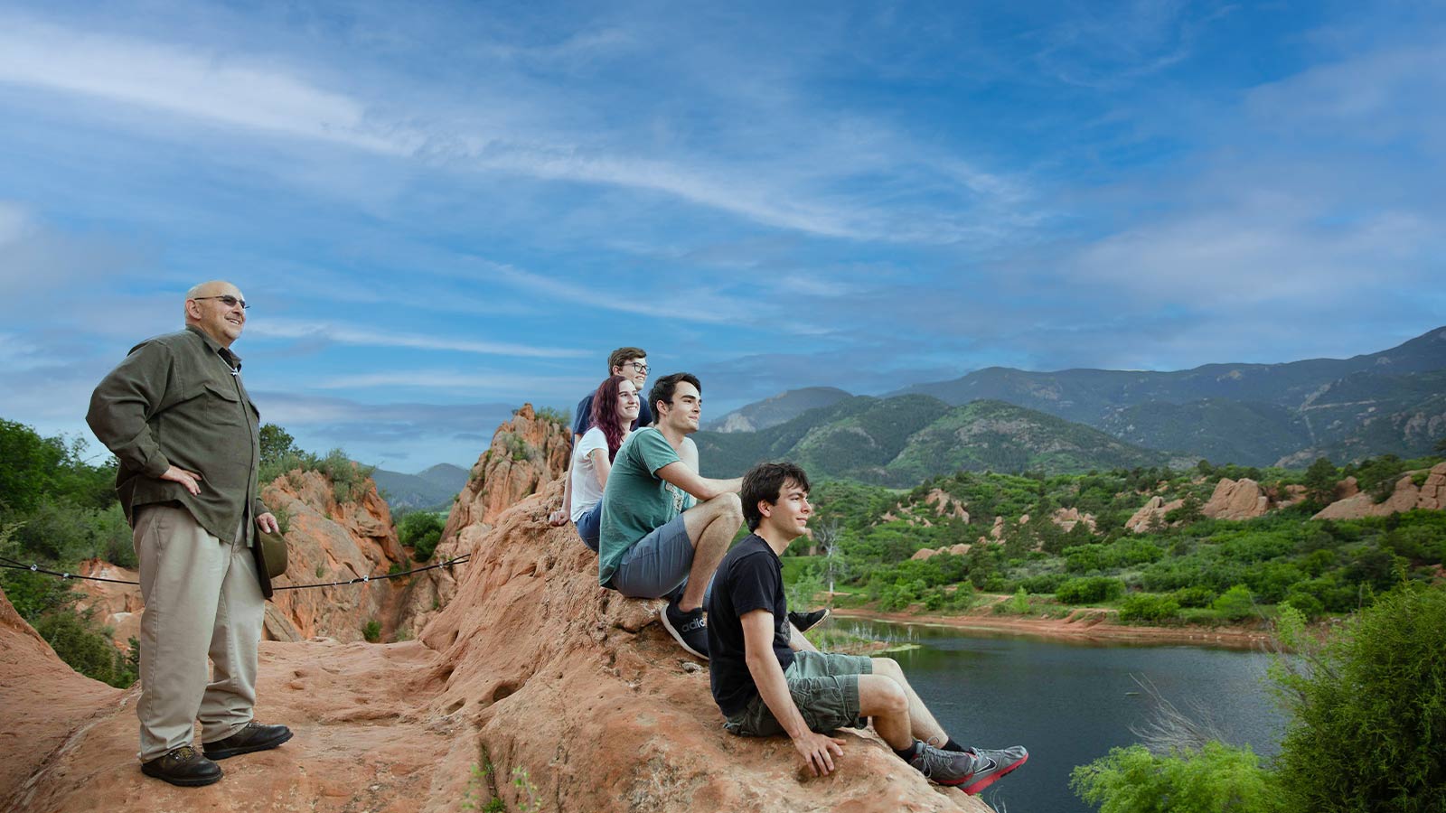 A group of four people sitting and standing on a red rock overlooking a scenic landscape with green hills and a lake.