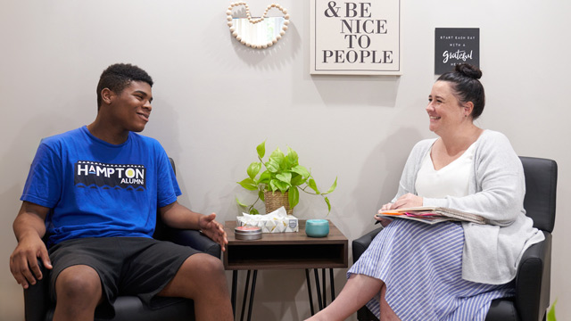 A young man and a therapist are smiling at each other, sitting in chairs in a cozy room with inspirational wall art.