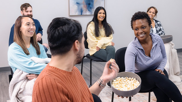A group of six people are enjoying a group therapy in a cozy room, with one person offering a bowl of popcorn.