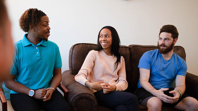 Three people sit on a brown sofa, sharing a moment of conversation with pleasant expressions.