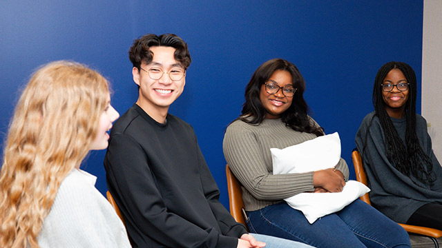 Four individuals are seated with smiles in group therapy against a deep blue wall in a casual setting.