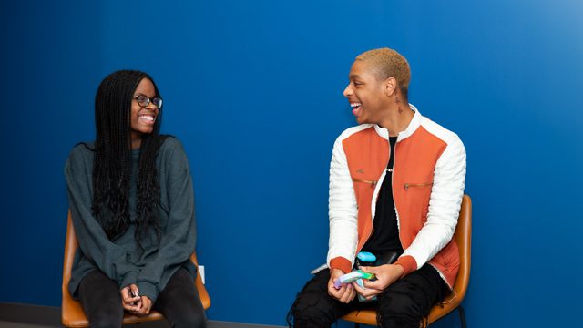 Two young adults are laughing and sitting on chairs in a room with a vibrant blue wall, enjoying a conversation.