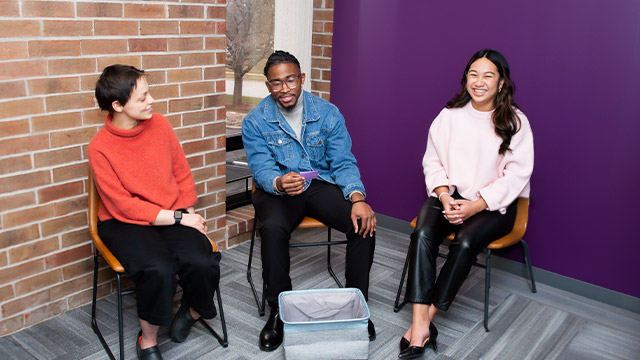 Three people sit in chairs, smiling and conversing, against a backdrop of a purple wall and exposed brick.