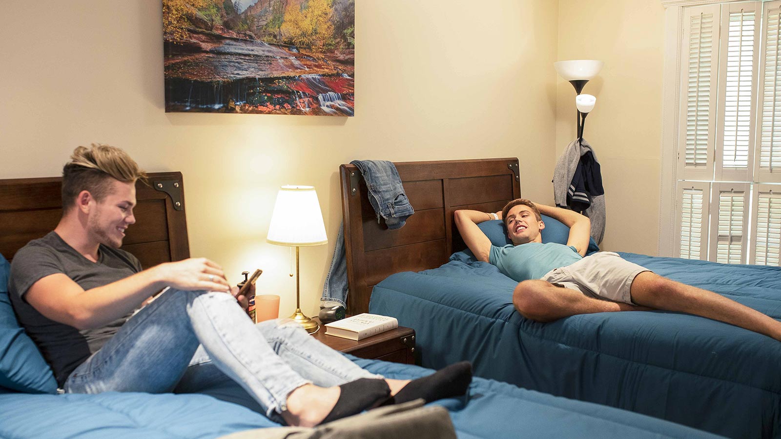Two young men are relaxing in a bedroom with one lying on his back on a bed, looking at his phone, and the other sitting with crossed legs on another bed, smiling while looking at his phone.