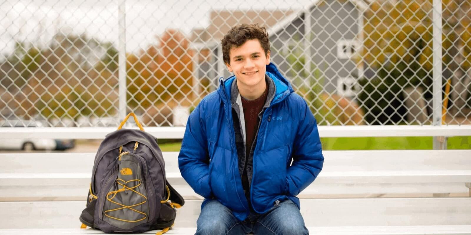 A teen boy smiling at the camera while sitting on a school bench