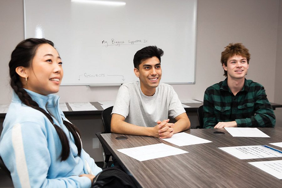 Three young adults smiling sitting at a table in a classroom, with a whiteboard in the background.