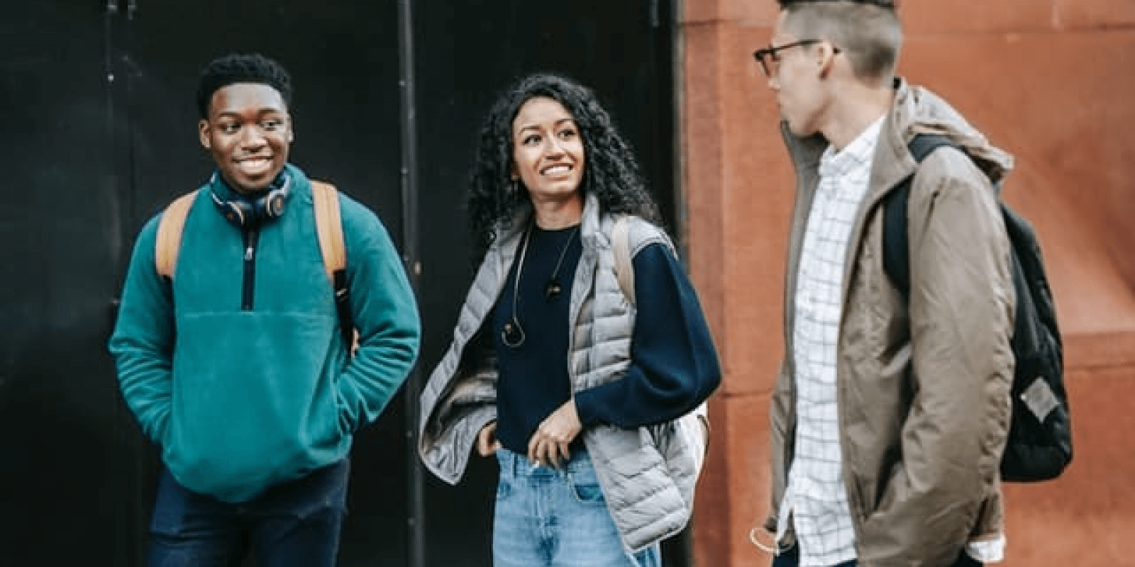 3 young adults wearing backpack walking down the street talking to each other