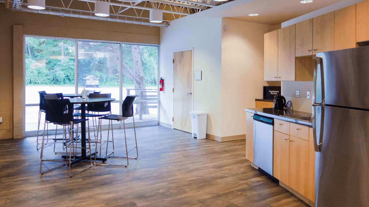 Sandstone Care's Reston Rehab Centers open kitchenette and dining area with high-top tables and chairs, modern appliances, and ample natural light coming through large windows, overlooking outdoor greenery