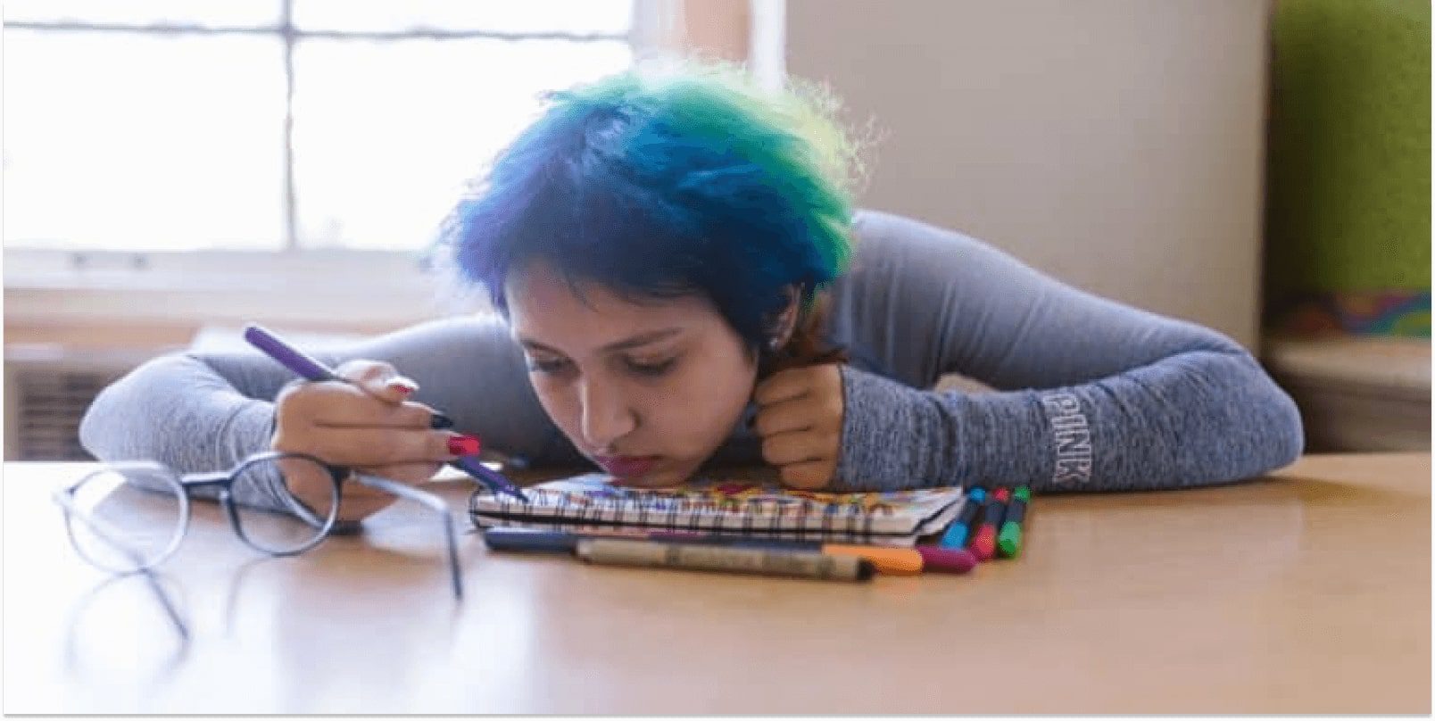 A blue hair teen putting her chin down to a drawing book on a table