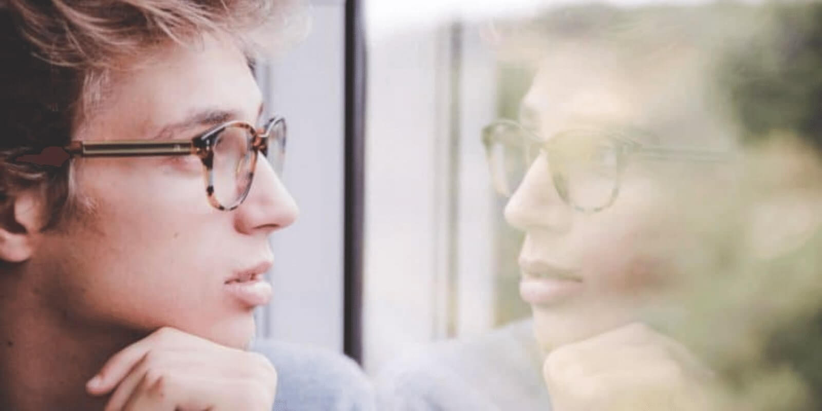 A teen boy with glasses looking at his reflection on a window