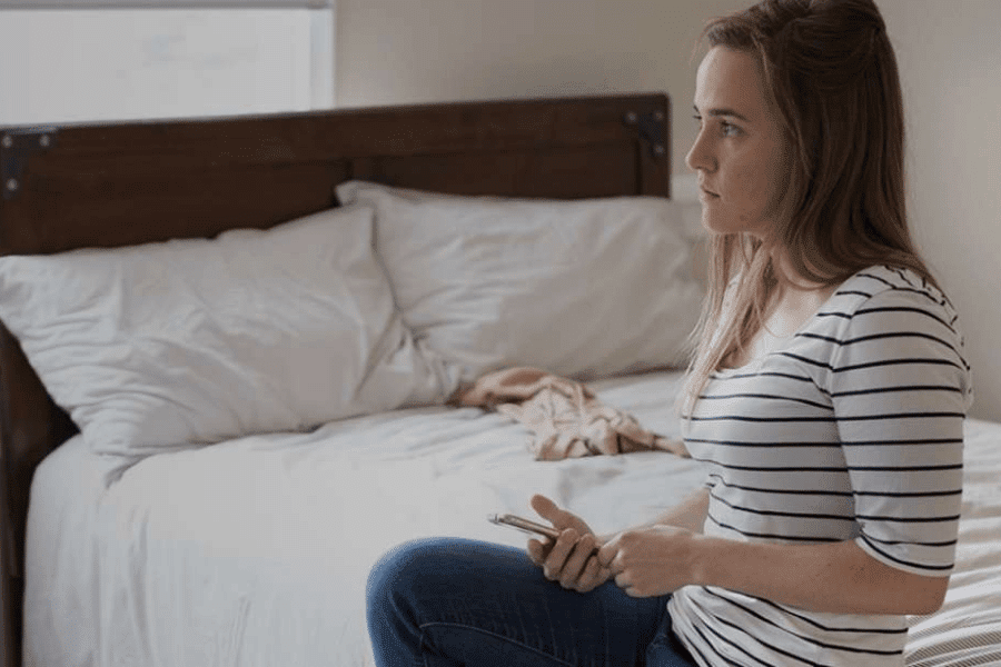 A young woman sitting on the bed looking away while holding a phone