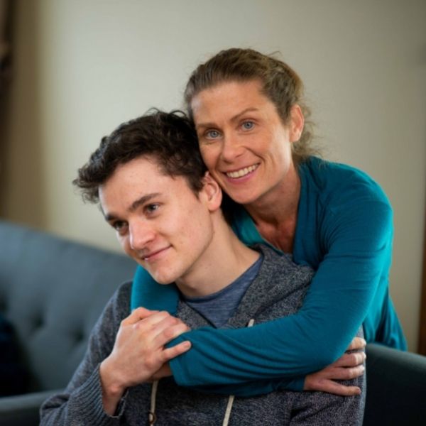 A mother hugging her teenage son while facing the camera.