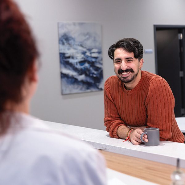 A man smiling and holding a coffee mug, seated at a white countertop having a conversation.