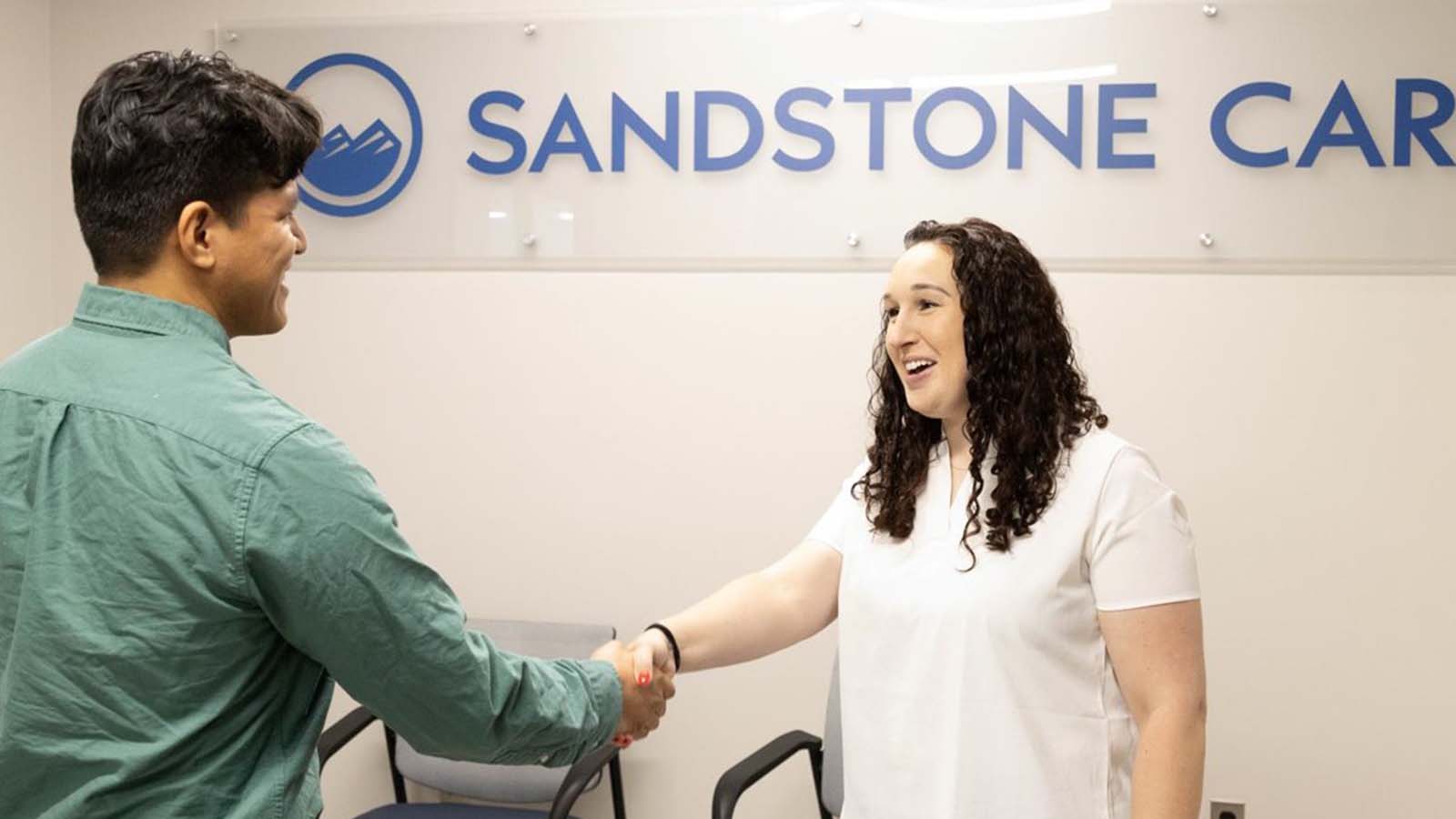Mental health therapist greeting and shaking hands with new patient in front of Sandstone Care logo.