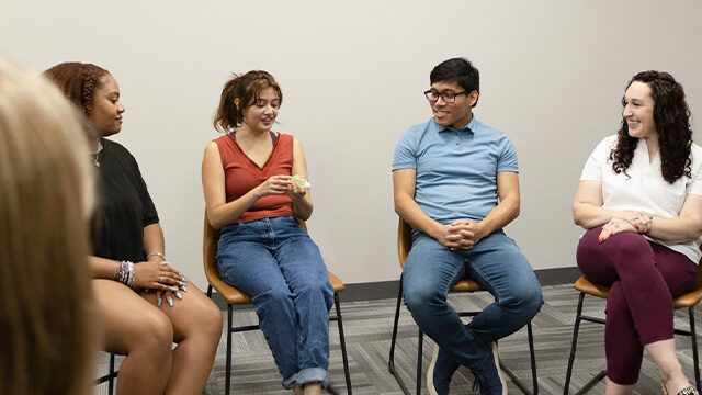 A diverse group of five young adults is seated in a circle, engaged in a discussion in a room with neutral tones.