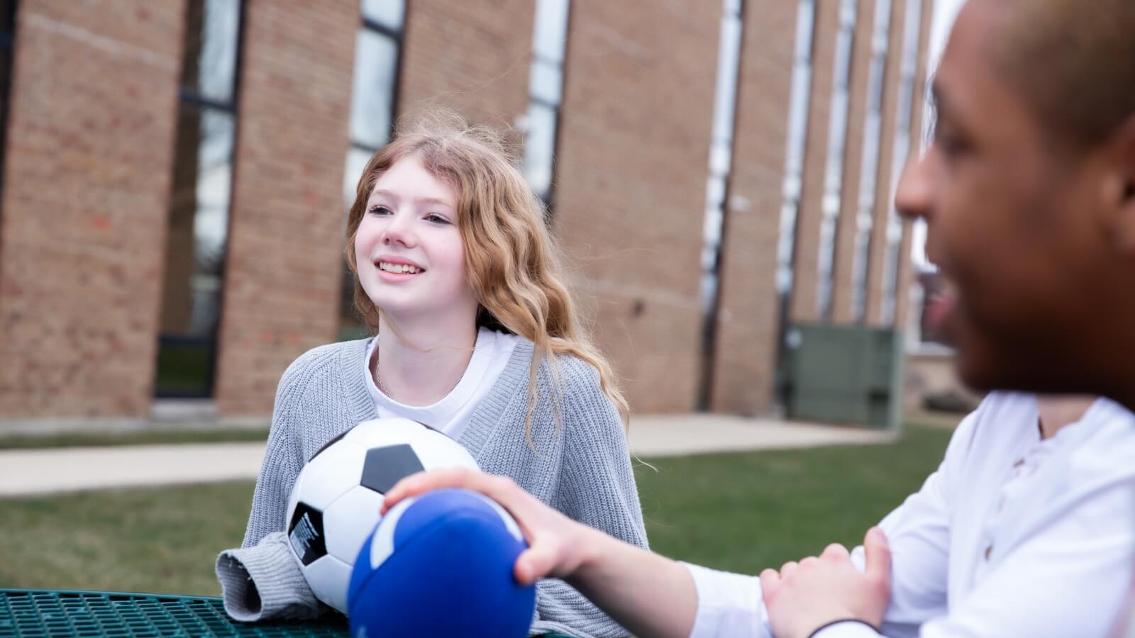 A young girl sitting at an outdoor table holding a soccer ball and engages in conversation with a person opposite her.